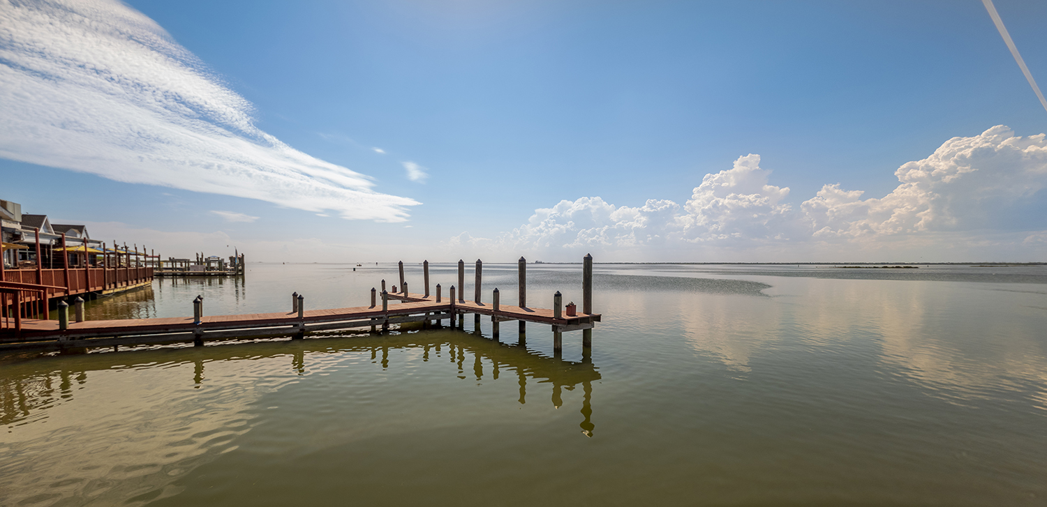 Snoopy's Pier extending over the calm waters of the Laguna Madre near Padre Island under a bright blue sky with reflected clouds.