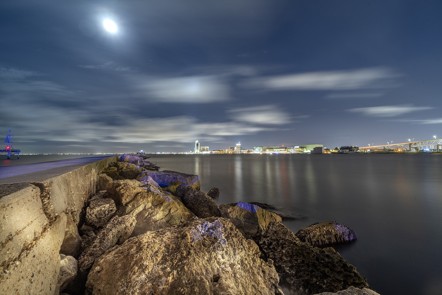 Night view across Corpus Christi Bay from the Bayfront seawall near the USS Lexington with moonlit clouds, calm water, and the downtown skyline in the distance.